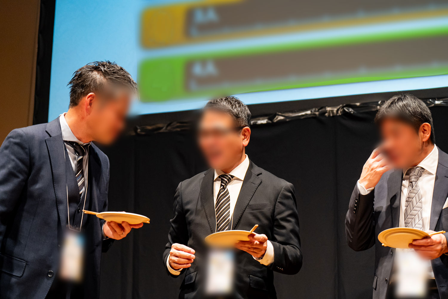 ステージ上で談笑しながら紙皿に乗っている食事を食べるスーツ姿の男性